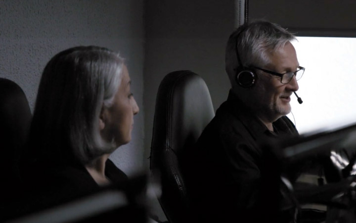A man with grey hair and glasses, wearing a head set speaks with a grey-haired woman